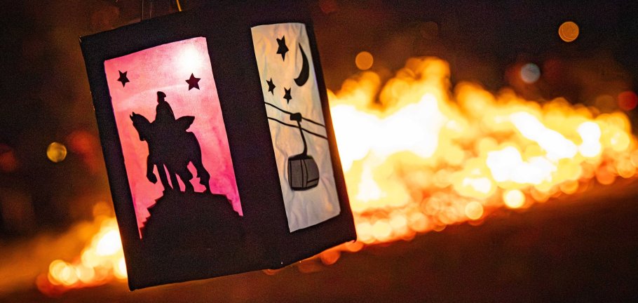 Eine Laterne leuchtet im Vordergrund, während im Hintergrund ein Feuer zu sehen ist.